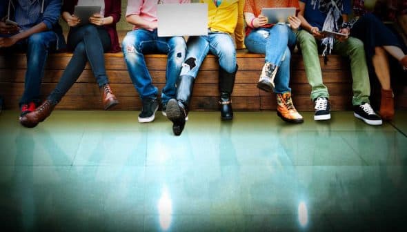 A group of individuals sitting on a bench, each using various electronic devices such as laptops and tablets. - Olive Oil Times