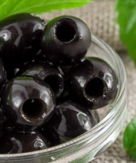 A close-up of black olives in a glass bowl with green leaves nearby. - Olive Oil Times