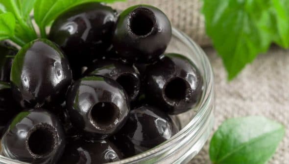 A close-up of black olives in a glass bowl with green leaves nearby. - Olive Oil Times