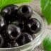A close-up of black olives in a glass bowl with green leaves nearby. - Olive Oil Times