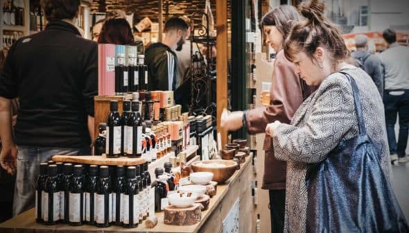 Various bottles of olive oil displayed on a wooden table at a market with people in the background. - Olive Oil Times