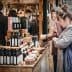 Various bottles of olive oil displayed on a wooden table at a market with people in the background. - Olive Oil Times