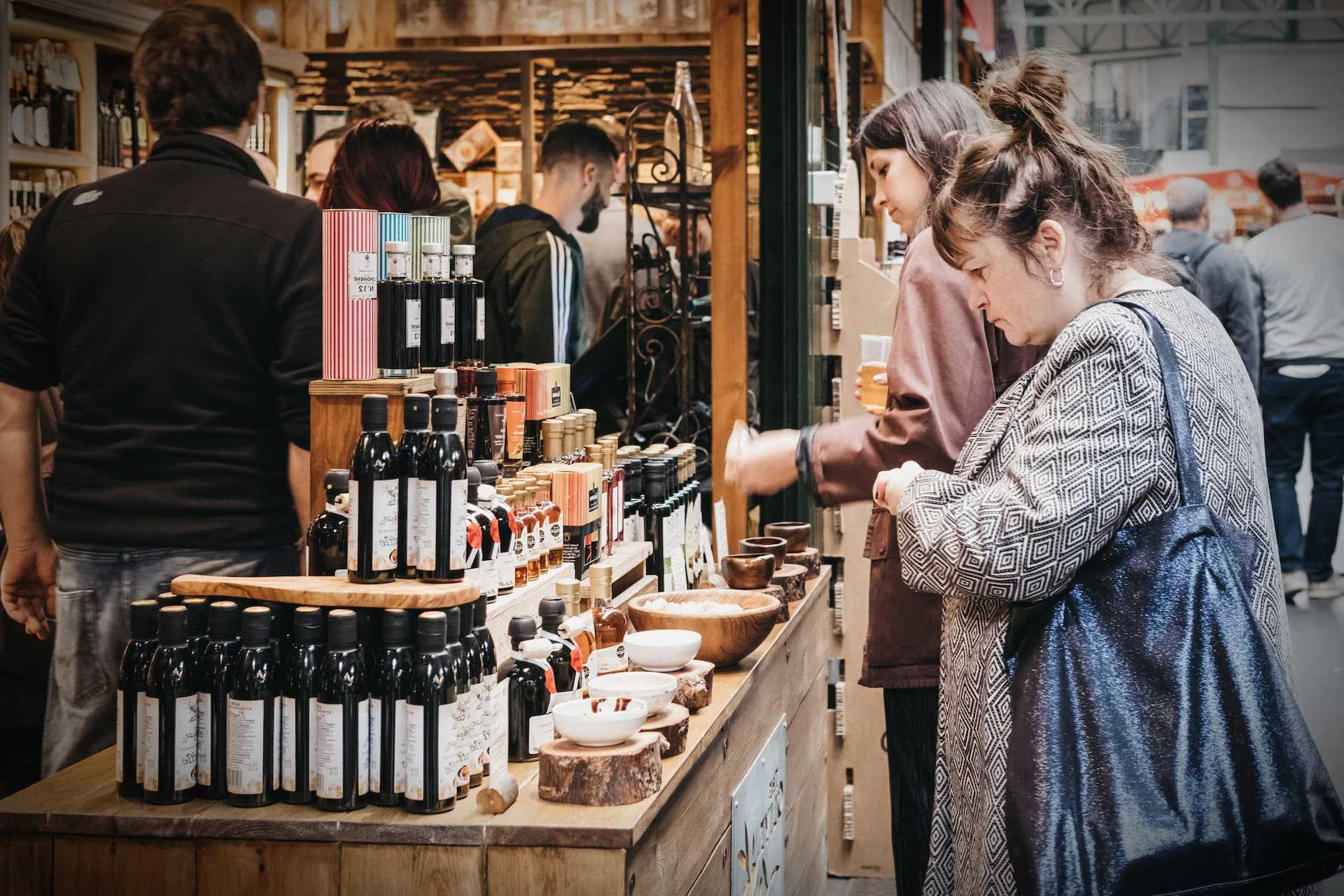 Various bottles of olive oil displayed on a wooden table at a market with people in the background. - Olive Oil Times