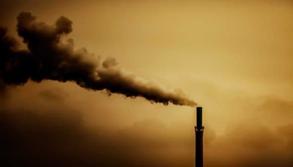 Smoke billowing from the top of an industrial smokestack against a cloudy sky. - Olive Oil Times