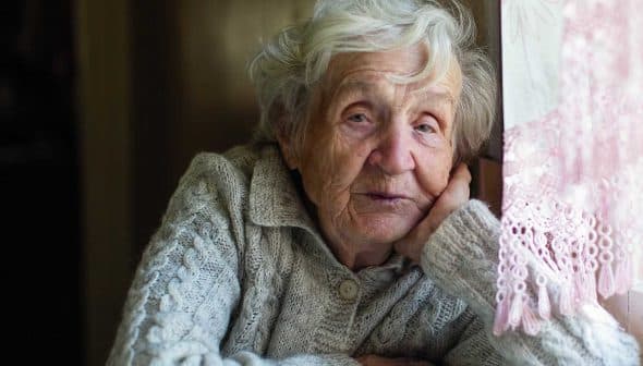Elderly woman with gray hair resting her head on her hand while sitting indoors in a sweater. - Olive Oil Times