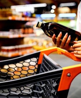 Individual holding a bottle of olive oil while standing next to a shopping cart in a store. - Olive Oil Times