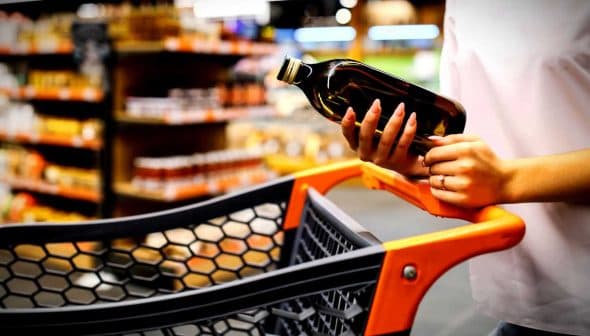 Individual holding a bottle of olive oil while standing next to a shopping cart in a store. - Olive Oil Times