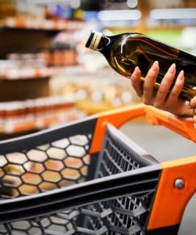 A person holding a bottle of olive oil while standing next to a shopping cart in a grocery store. - Olive Oil Times