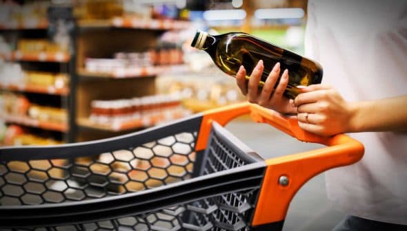 A person holding a bottle of olive oil while standing next to a shopping cart in a grocery store. - Olive Oil Times