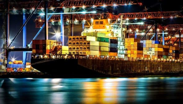 Cargo ship docked at a port, loaded with colorful shipping containers at night. - Olive Oil Times