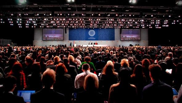 A large audience gathered in a convention center for a conference event with screens displaying presentations. - Olive Oil Times