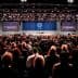 A large audience gathered in a convention center for a conference event with screens displaying presentations. - Olive Oil Times