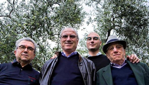Four men standing together outdoors among olive trees, all looking at the camera. - Olive Oil Times