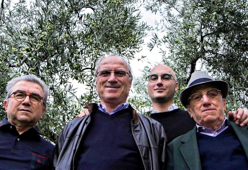 Four men standing together outdoors among olive trees, all looking at the camera. - Olive Oil Times