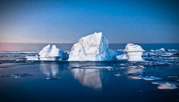 Icebergs floating in calm waters with a blue sky in the background during twilight. - Olive Oil Times
