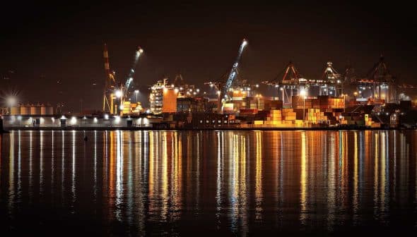 Night view of a port with illuminated shipping containers and cranes reflecting on the water. - Olive Oil Times