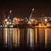 Night view of a port with illuminated shipping containers and cranes reflecting on the water. - Olive Oil Times