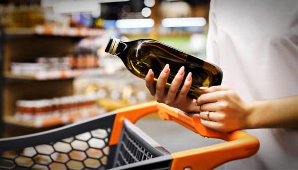 Individual holding a dark glass bottle of olive oil while standing next to a shopping cart. - Olive Oil Times