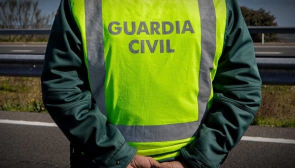A Guardia Civil officer wearing a high-visibility vest stands with hands behind back on a roadside. - Olive Oil Times