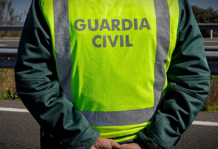 A Guardia Civil officer wearing a high-visibility vest stands with hands behind back on a roadside. - Olive Oil Times