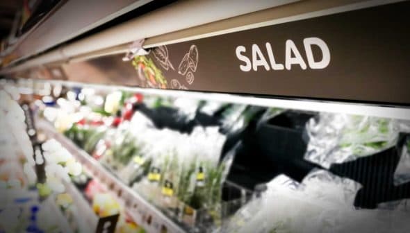 Sign labeled 'SALAD' in a grocery store aisle with various salad ingredients visible. - Olive Oil Times