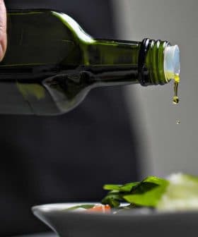 A person pouring olive oil from a dark green bottle onto a fresh salad in a white bowl. - Olive Oil Times