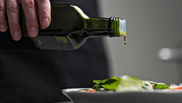 A person pouring olive oil from a dark green bottle onto a fresh salad in a white bowl. - Olive Oil Times
