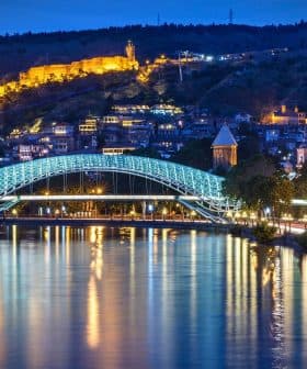 Illuminated bridge over a river with a cityscape and hillside in the background at night. - Olive Oil Times