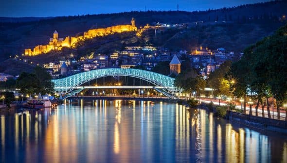 Illuminated bridge over a river with a cityscape and hillside in the background at night. - Olive Oil Times