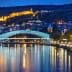 Illuminated bridge over a river with a cityscape and hillside in the background at night. - Olive Oil Times
