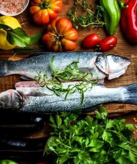 Two whole fish placed on a wooden board surrounded by various vegetables and herbs. - Olive Oil Times