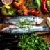 Two whole fish placed on a wooden board surrounded by various vegetables and herbs. - Olive Oil Times