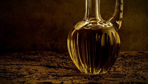 A glass decanter filled with olive oil, featuring a ribbed design and a curved spout. - Olive Oil Times