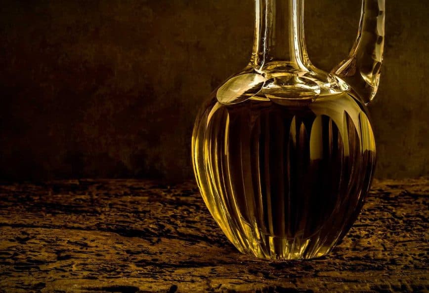 A glass decanter filled with olive oil, featuring a ribbed design and a curved spout. - Olive Oil Times