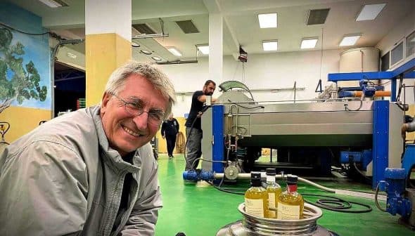 A man smiling in an olive oil processing facility with machinery and bottles of olive oil in the foreground. - Olive Oil Times