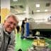 A man smiling in an olive oil processing facility with machinery and bottles of olive oil in the foreground. - Olive Oil Times