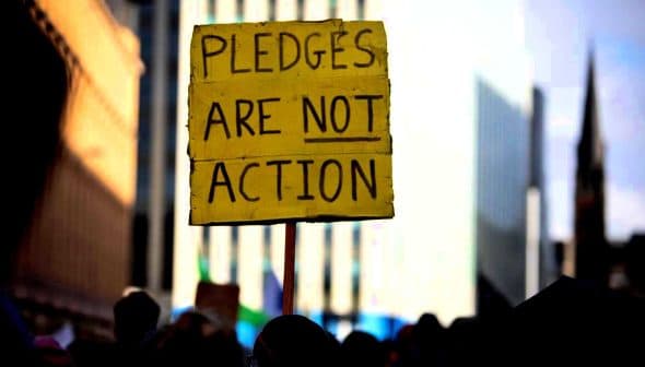 Yellow sign at a protest reading 'Pledges Are Not Action' held by an individual in a crowd. - Olive Oil Times