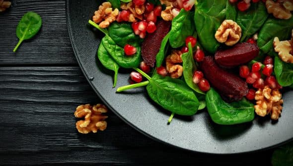A black plate filled with spinach leaves, pomegranate seeds, and walnuts arranged together. - Olive Oil Times