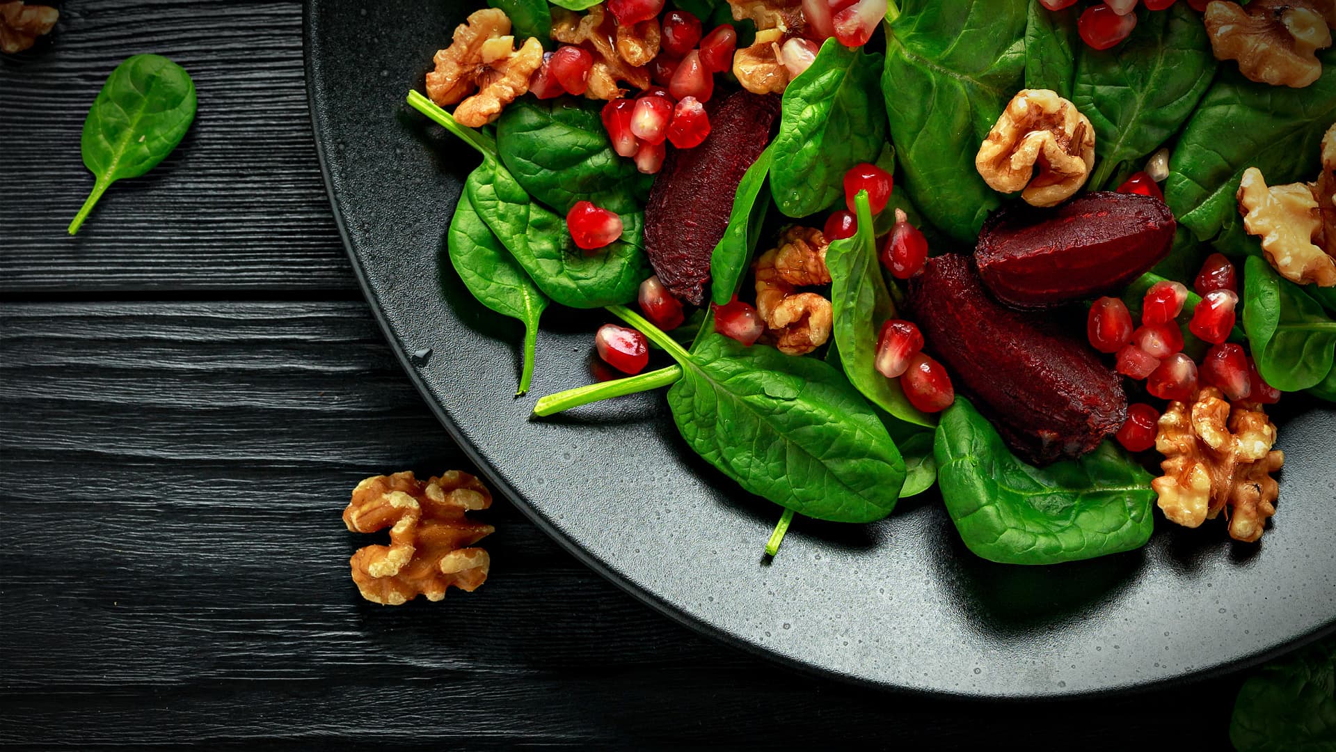 A black plate filled with spinach leaves, pomegranate seeds, and walnuts arranged together. - Olive Oil Times