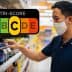 Man wearing a mask examining beverage labels on a supermarket shelf with a Nutri-Score graphic in the background. - Olive Oil Times