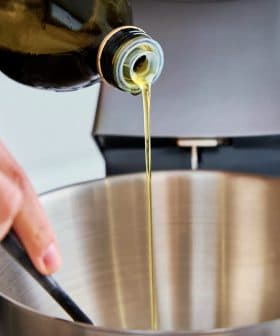 A person pouring olive oil from a bottle into a stainless steel mixing bowl. - Olive Oil Times