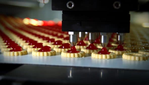 Automated machine dispensing red filling onto cookie bases in a production line. - Olive Oil Times