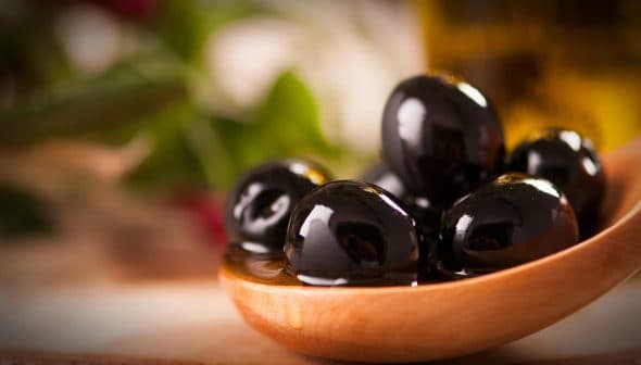 A wooden spoon holding several black olives in olive oil, with a blurred background. - Olive Oil Times