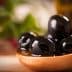 A wooden spoon holding several black olives in olive oil, with a blurred background. - Olive Oil Times