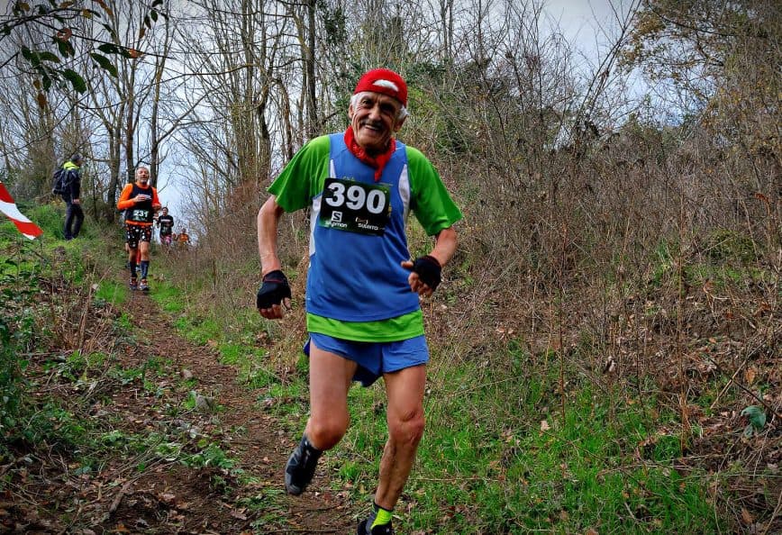 Two trail runners participating in a race on a wooded path, one wearing a green and blue outfit with a race number. - Olive Oil Times
