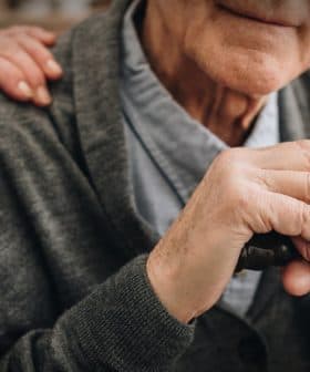 Close-up of an elderly man's hands resting on a cane, with a hand on his shoulder. - Olive Oil Times