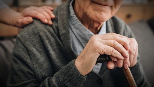 Close-up of an elderly man's hands resting on a cane, with a hand on his shoulder. - Olive Oil Times