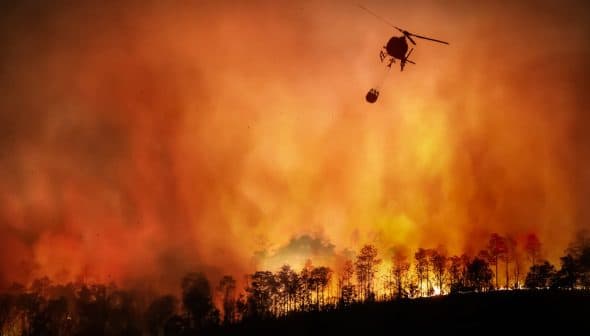 Helicopter dropping water on a large wildfire with flames and smoke in the background. - Olive Oil Times