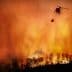 Helicopter dropping water on a large wildfire with flames and smoke in the background. - Olive Oil Times