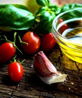 Cherry tomatoes, garlic, fresh basil, and a small bowl of olive oil on a wooden surface. - Olive Oil Times